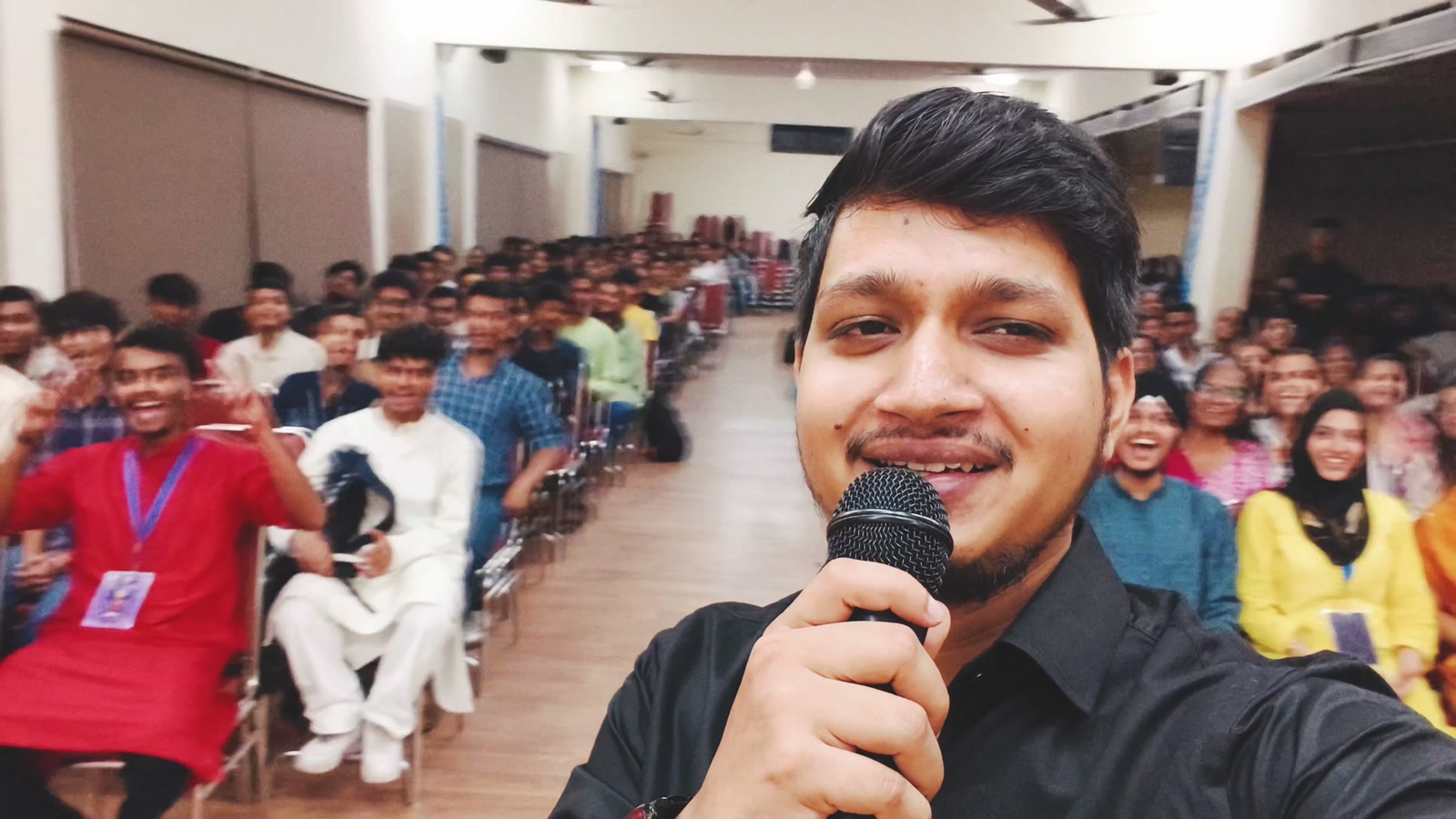 Ali Mustufa Shaikh holding a microphone and filming himself with a seated audience smiling and waving behind him.
