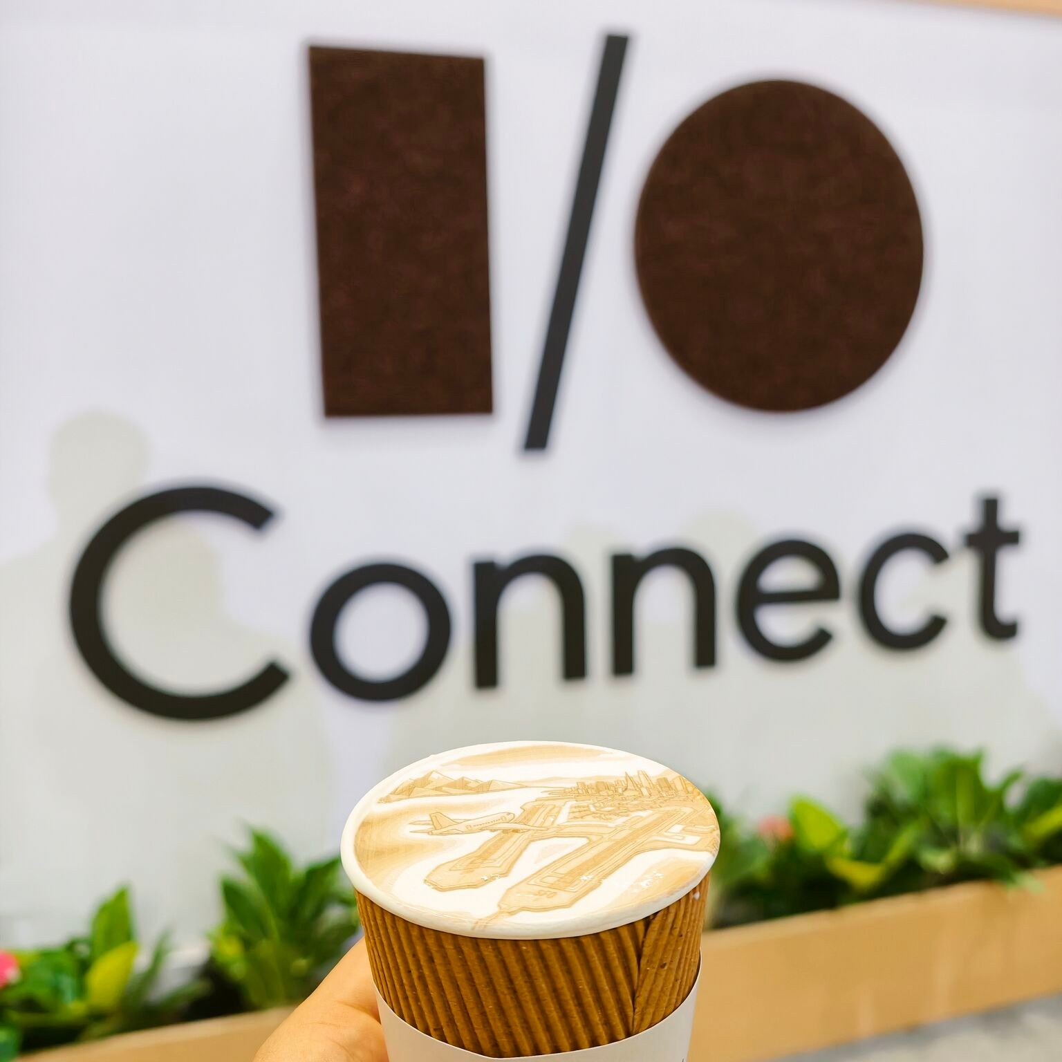 A takeaway cup topped with latte art depicting an airport and city skyline, held in front of an I/O Connect sign.
