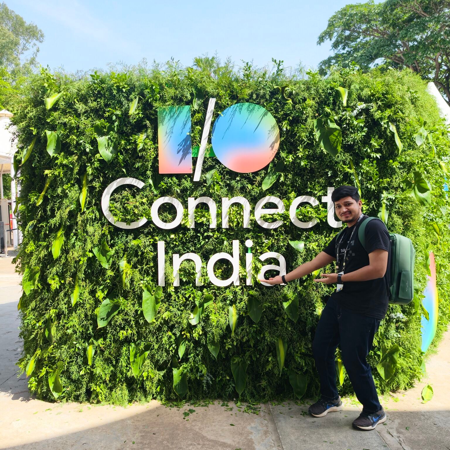 Ali Mustufa Shaikh stands beside a large Google I/O Connect India sign mounted on a lush green wall.